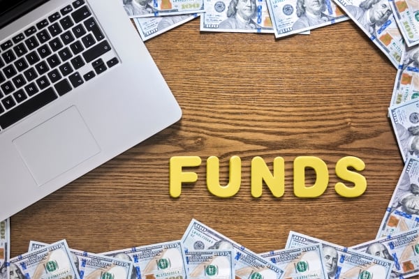 fundsforNGOs - 2026-06-02T142934590 - fundsforNGOs Laptop on a wooden desk with scattered dollar bills and yellow letters spelling FUNDS nearby.