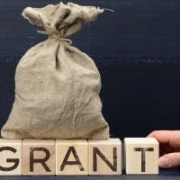 Burlap money bag on top of wooden blocks spelling GRANT with a hand nearby, symbolizing funding