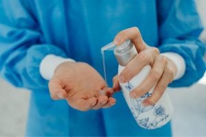 Person in a blue protective gown pours hand sanitizer into their palm from a white bottle with a floral design.