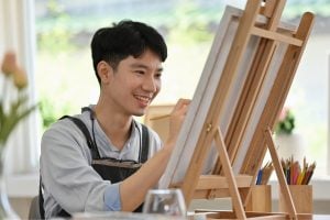 Young man painting on a wooden easel in a bright studio, smiling as he works with a brush and canvas near colorful pencils and a glass on the table