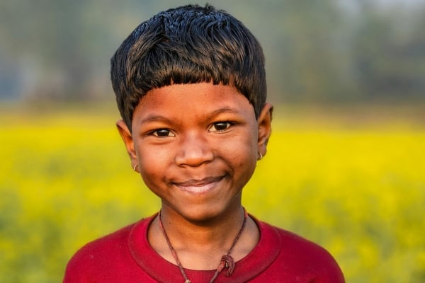 fundsforNGOs - 2026-06-02T150724632 - fundsforNGOs Smiling young boy with short black hair wearing a red shirt, standing in a field of yellow flowers.