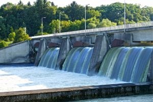 Hydroelectric dam with multiple sluice gates releasing water into the reservoir, backed by green trees and a roadway above the dam.
