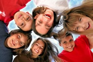 Six smiling children form a circle, looking down at the camera outdoors against a blue sky.