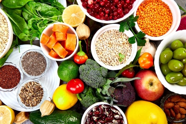 Colorful assortment of fresh vegetables, fruits, and grains in bowls and on a table. Includes leafy greens, citrus, berries, legumes, and seeds.
