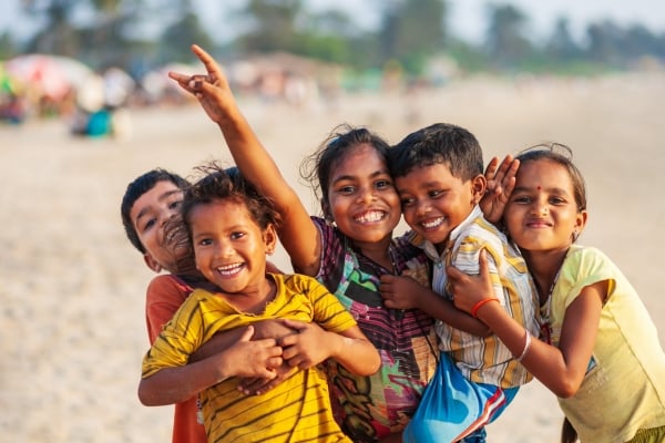 Five children on a sunny beach hug and smile at the camera, with blurred beachgoers in the background.