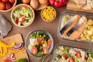 Assorted prepared meals on a table: salmon fillets, mixed salads, corn, peppers, and onions in bowls and containers.