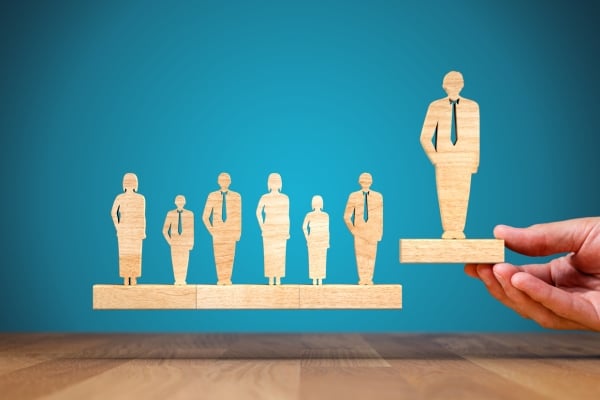 Hand places a wooden figure on a pedestal, elevating a leader above a row of other wooden figures against a blue background.