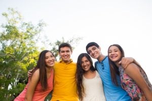 Five young friends with arms around each other pose for a sunny outdoor photo, smiling at the camera.