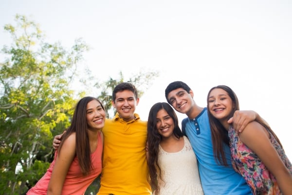 Five young friends with arms around each other pose for a sunny outdoor photo, smiling at the camera.