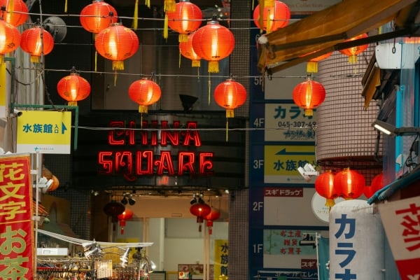 Red lanterns hang across a busy street leading to a storefront with a bright neon 'CHINA SQUARE' sign. Japanese signs line the buildings on both sides.