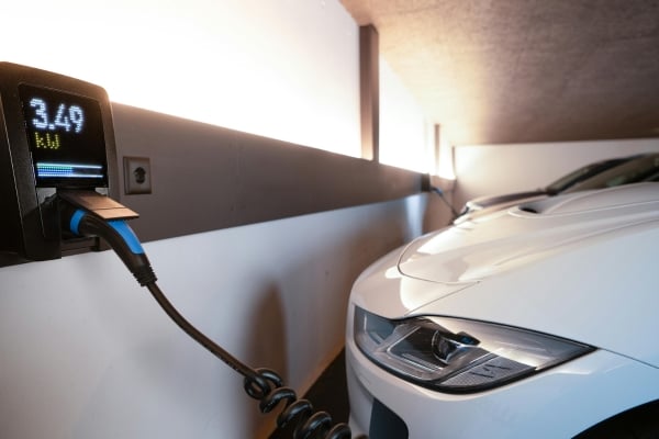Electric car charging in a dim garage; wall-mounted charger displays 3.49 kW while a coiled cable plugs into the white vehicle.
