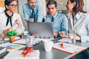 Four professionals gathered around a laptop in a meeting, reviewing documents and colorful charts on the table, in a bright office.