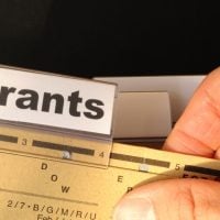 fundsforNGOs - 2026-06-04T153003556 - fundsforNGOs Close-up of a label reading 'Grants' being slid into a labeler above a brass ruler filled with marks and holes on a desk surface, with a finger nearby.