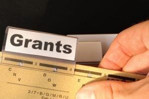 Close-up of a label reading 'Grants' being slid into a labeler above a brass ruler filled with marks and holes on a desk surface, with a finger nearby.