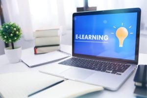 Laptop displaying an e-learning concept with a bright light bulb on the screen, on a desk with books and a notebook nearby.