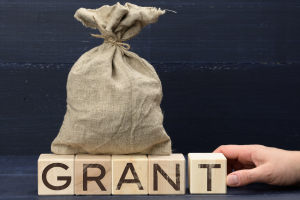 A burlap money bag sits on wooden blocks that spell GRANT, with a hand nearby, suggesting funding or grant support.