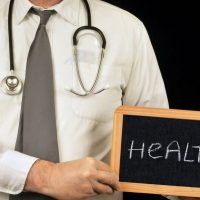 health - fundsforNGOs Doctor with stethoscope holding a small chalkboard that reads HEALTH, foregrounded against a dark background to convey health care theme.
