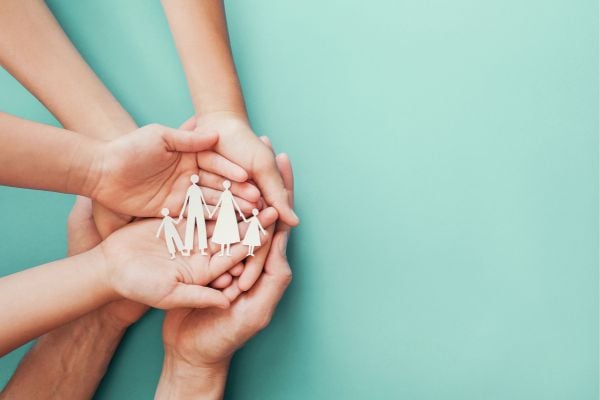 health 7 - fundsforNGOs Hands of adults and children cupped together around a white paper cutout of a family, symbolizing support and unity on a teal background.