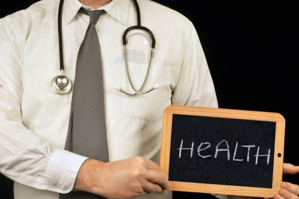 Doctor with stethoscope holding a small chalkboard that reads HEALTH, foregrounded against a dark background to convey health care theme.