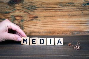 A hand places wooden letter blocks spelling MEDIA on a dark wooden table, with a small copper clip nearby.