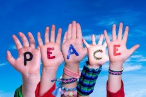Raised hands with colorful letters spelling 'PEACE' against a bright blue sky, suggesting a message of unity by kids/young people.