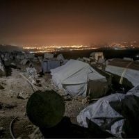 refugees - fundsforNGOs Nighttime campsite on a rocky hillside with beige tents and scattered gear, city lights glowing on the horizon.