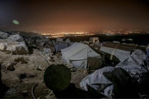 Nighttime campsite on a rocky hillside with beige tents and scattered gear, city lights glowing on the horizon.