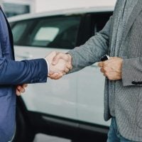 research 4 - fundsforNGOs Two men shake hands in front of a white car, signaling a business deal or agreement outside a dealership or showroom.