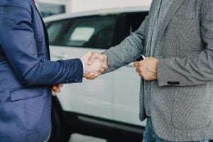 Two men shake hands in front of a white car, signaling a business deal or agreement outside a dealership or showroom.