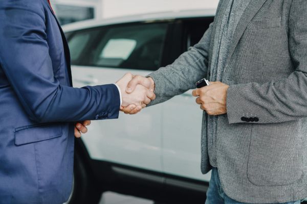 research 4 - fundsforNGOs Two men shake hands in front of a white car, signaling a business deal or agreement outside a dealership or showroom.