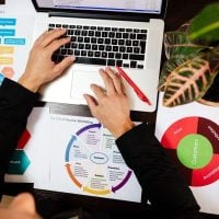 research 6 - fundsforNGOs Overhead view of a person typing on a laptop at a desk with printed marketing diagrams and a potted plant nearby, suggesting a marketing analysis session.