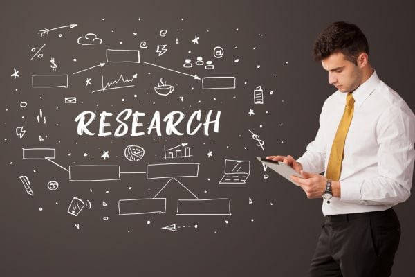 research 7 - fundsforNGOs Businessman in a white shirt and yellow tie uses a tablet, beside a chalkboard filled with research doodles and the word RESEARCH.