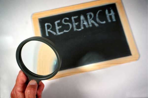 Person holds a magnifying glass up to a chalkboard that reads 'RESEARCH'.