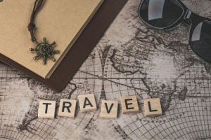 Scrabble tiles spell 'TRAVEL' on a vintage map with a notebook, sunglasses, and a compass pendant.