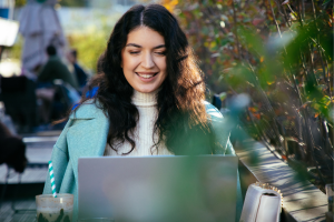 Young woman studying on a laptop at an outdoor cafe, smiling as she works with greenery around her.