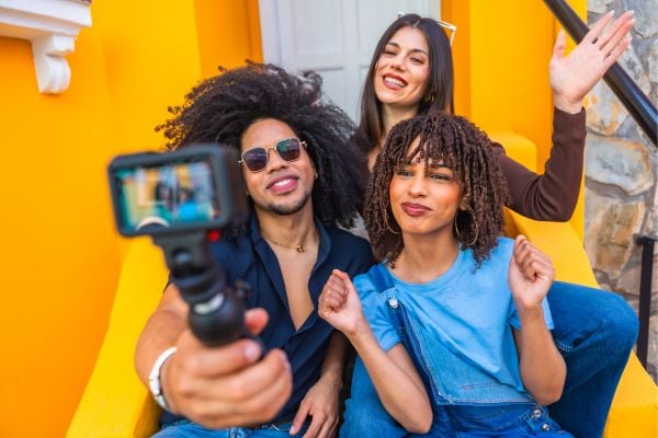 youth 2 - fundsforNGOs Three friends posing for a selfie on bright yellow steps, with one person holding a camera on a selfie stick/holder.