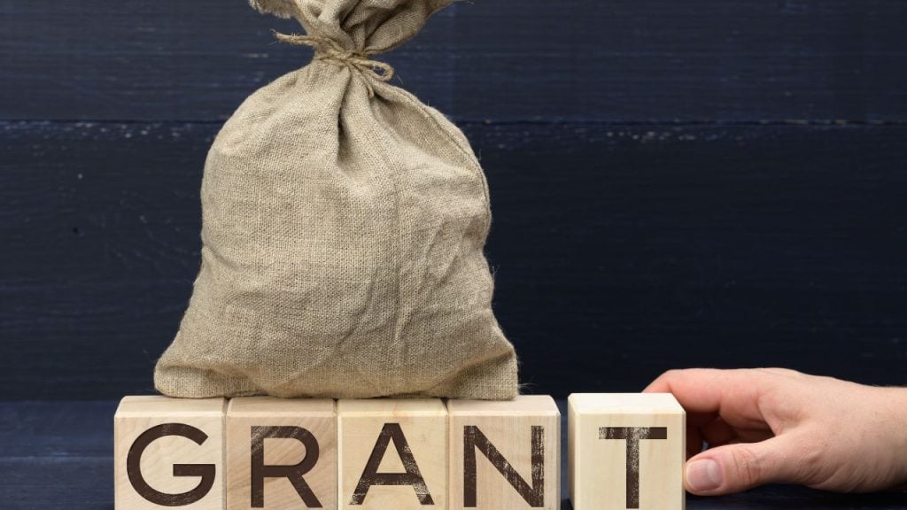 education and sports for children - fundsforNGOs A burlap sack sits on wooden blocks spelling GRANT, with a hand touching the final block, suggesting grant funds or approval.