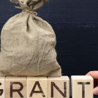 education and sports for children - fundsforNGOs A burlap sack sits on wooden blocks spelling GRANT, with a hand touching the final block, suggesting grant funds or approval.