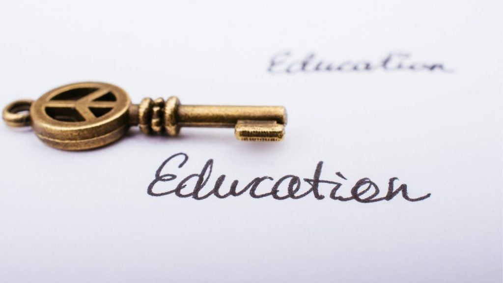 education and sports for children - fundsforNGOs Brass key with a peace-sign bow lies on a white page printed with the word 'Education' in cursive, symbolizing education as a key to peace.